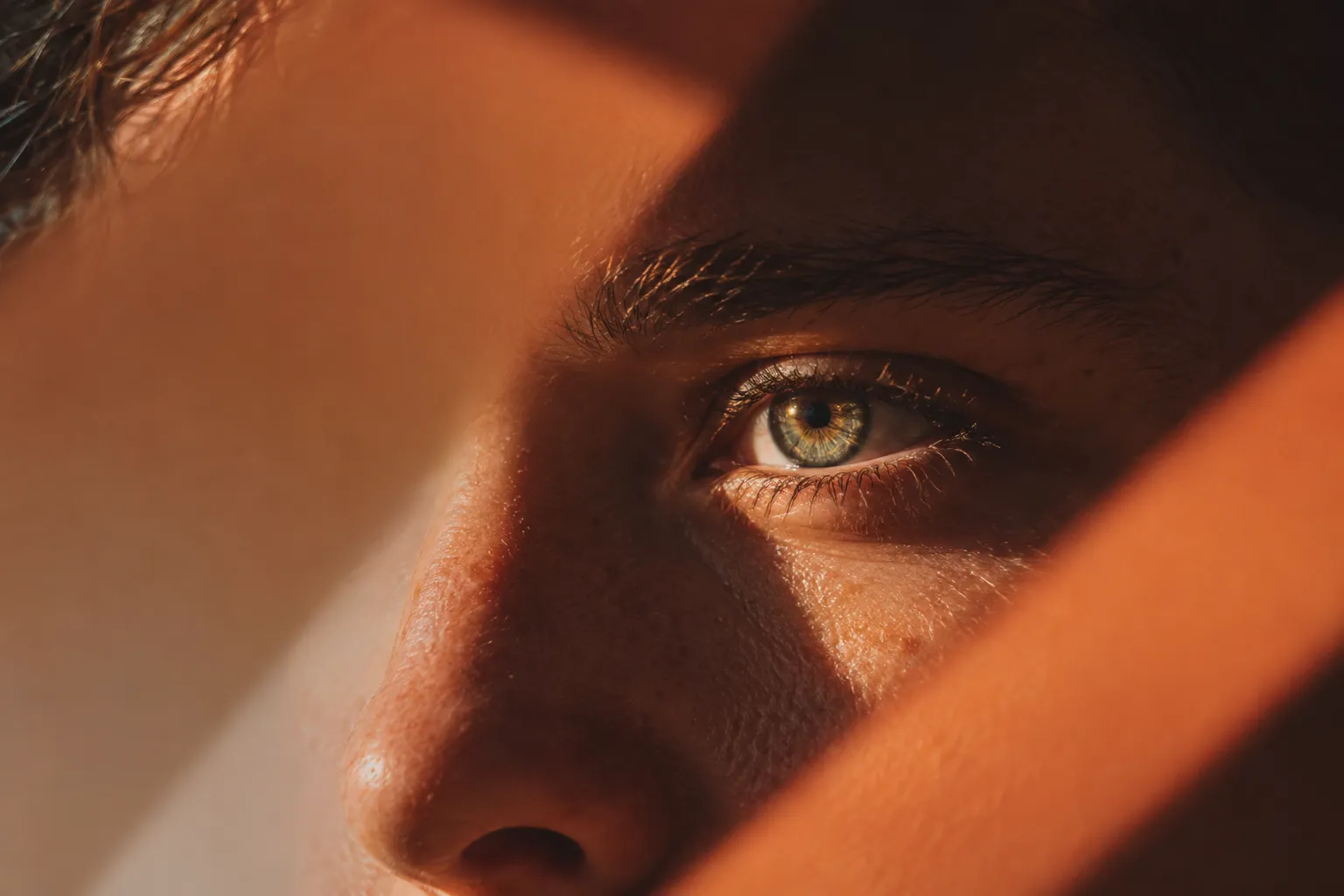 Extreme close crop of a face lit through hard diagonal bands of warm light, one green-hazel eye near the centre of the frame