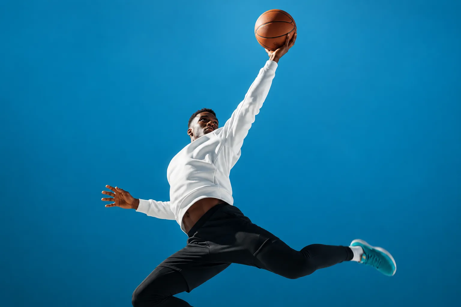 Athlete in a white long-sleeve top and black trousers leaping diagonally with one arm stretched up, palm under a basketball, against a seamless cerulean studio backdrop
