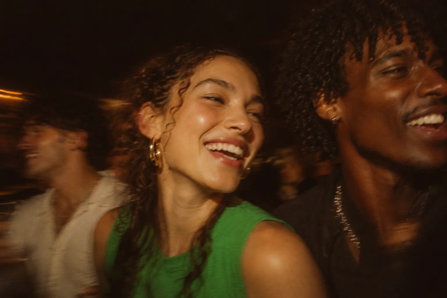 Three friends laughing together at a night party, caught in warm on-camera flash with motion blur